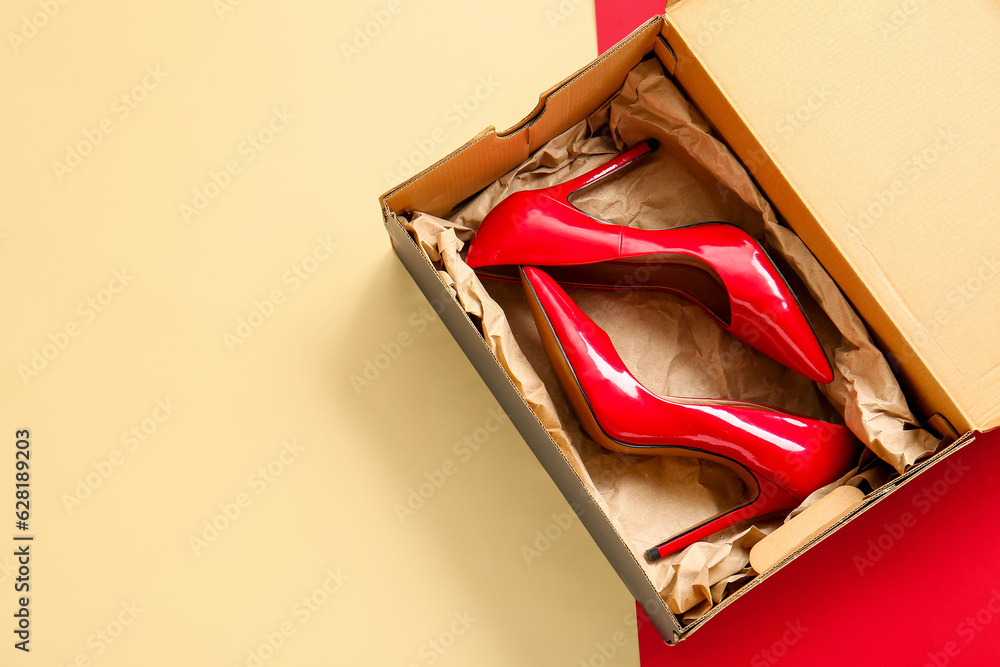 Cardboard box with high heeled shoes on color background