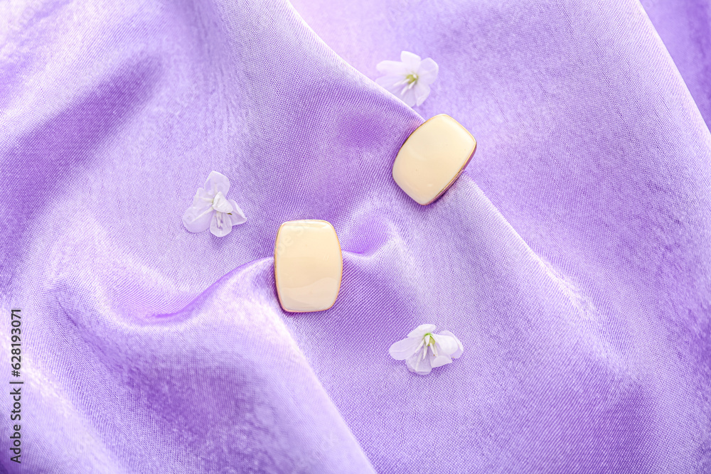 Stylish earrings with flowers on violet fabric background, closeup