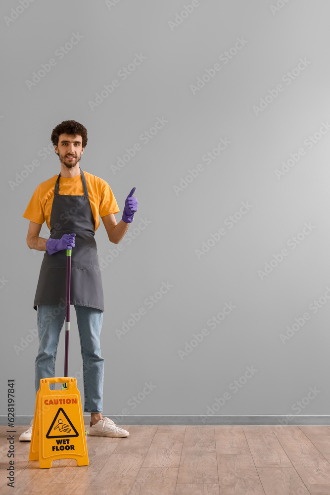 Young man with mop and caution sign showing thumb-up near grey wall