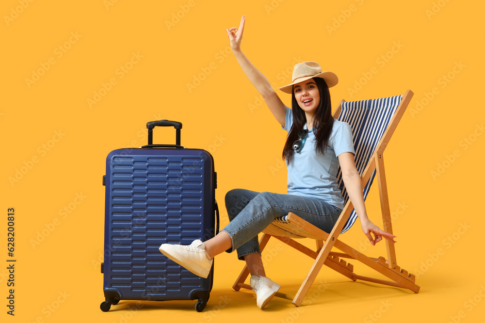 Young woman with suitcase in deck chair showing victory gesture on yellow background