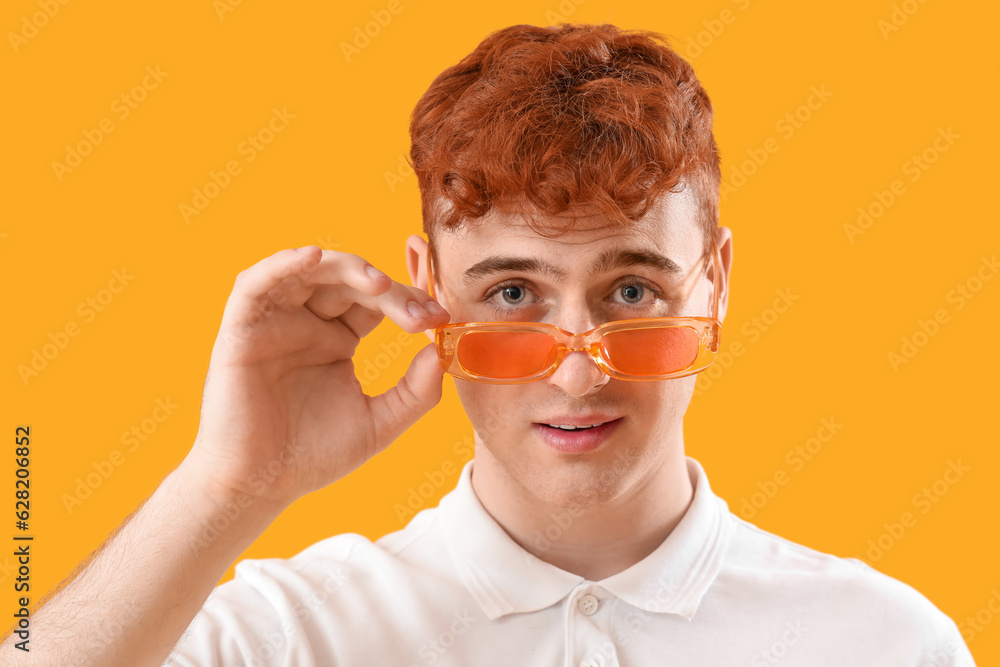 Young redhead man in stylish sunglasses on yellow background, closeup