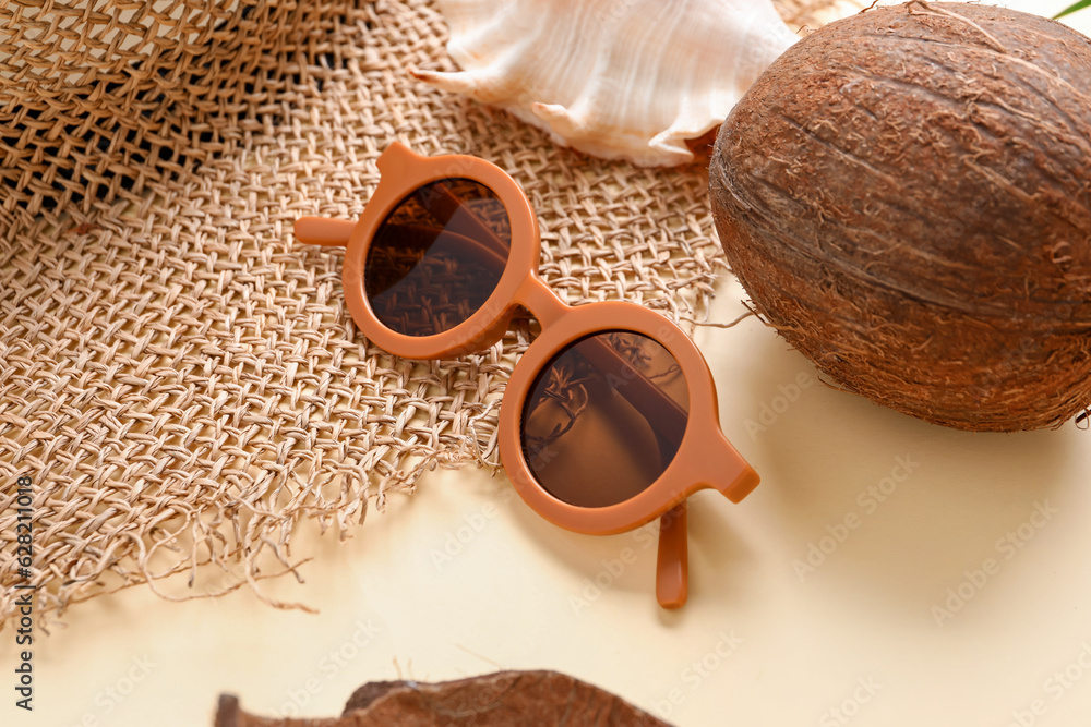 Stylish sunglasses with wicker hat and coconut on pale yellow background