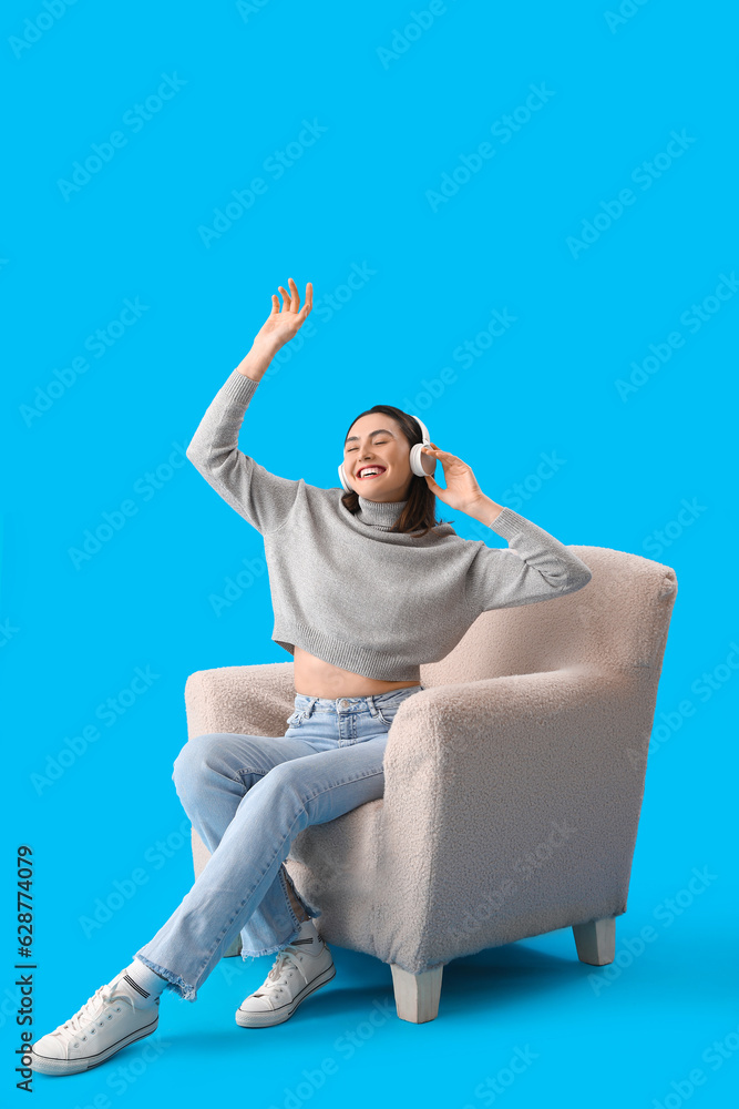 Young woman with headphones sitting in soft armchair on blue background