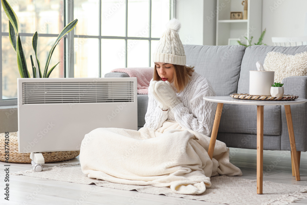 Frozen young woman in winter clothes warming near radiator at home