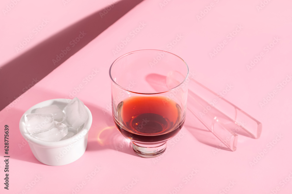 Glass of rum, bowl with ice cubes and tongs on pink background