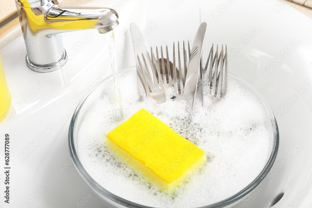 Bowl with cutlery and cleaning sponge in sink, closeup