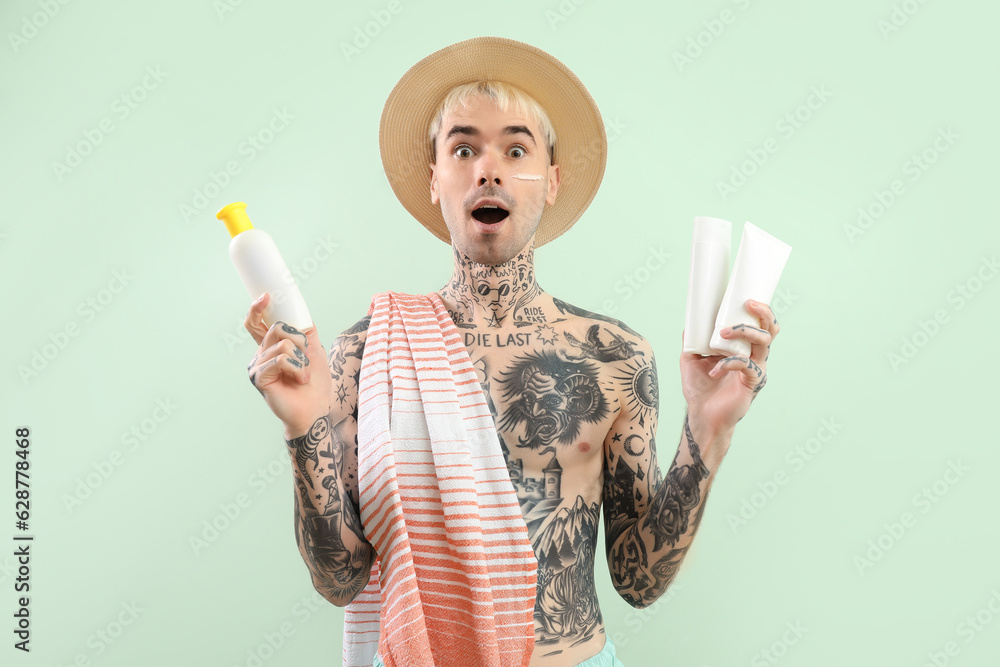 Surprised tattooed man with sunscreen creams on light green background