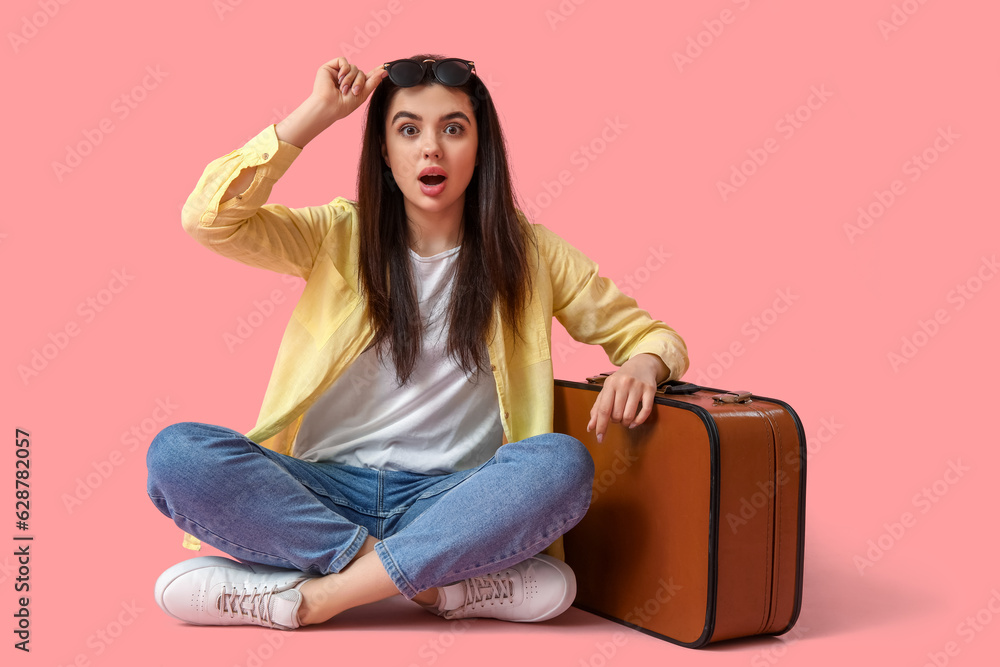 Surprised young woman with suitcase sitting on pink background