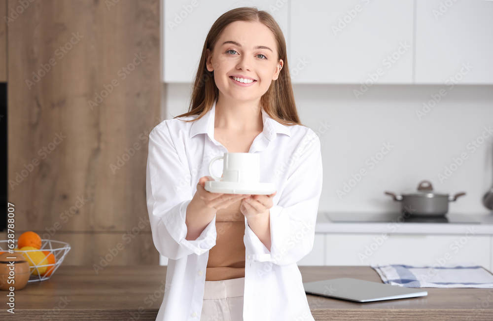 Happy young woman holding saucer with cup of coffee in modern kitchen