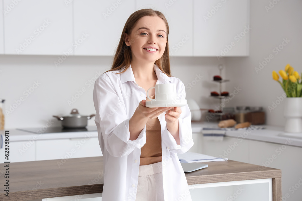 Happy young woman holding saucer with cup of coffee in modern kitchen