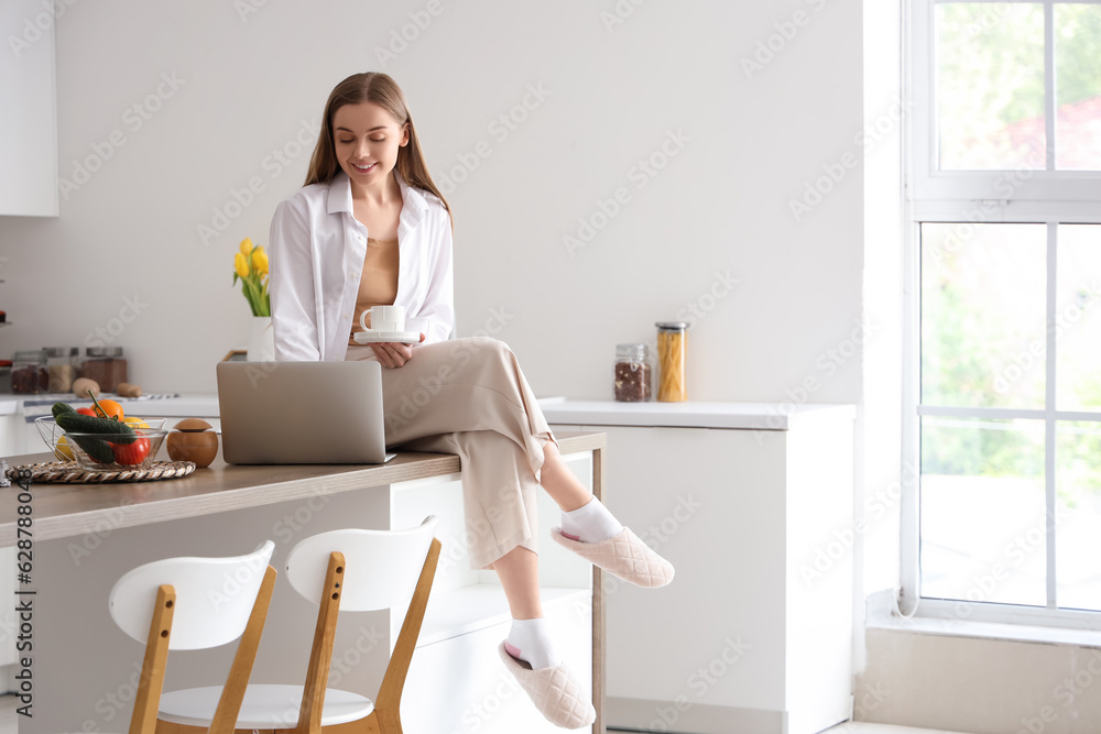 Happy young woman holding cup of coffee and sitting on table with modern laptop in light kitchen