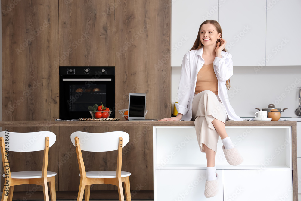 Happy young woman sitting on table with modern laptop in light kitchen