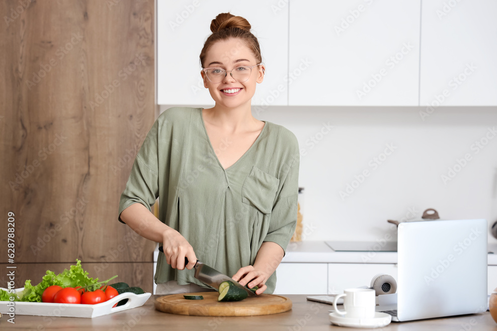 Beautiful young woman cutting cucumber at table with modern laptop in light kitchen