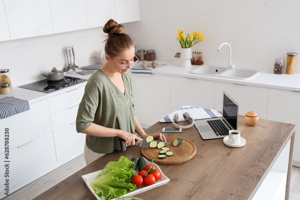 Beautiful young woman cutting cucumber at table with modern laptop in light kitchen