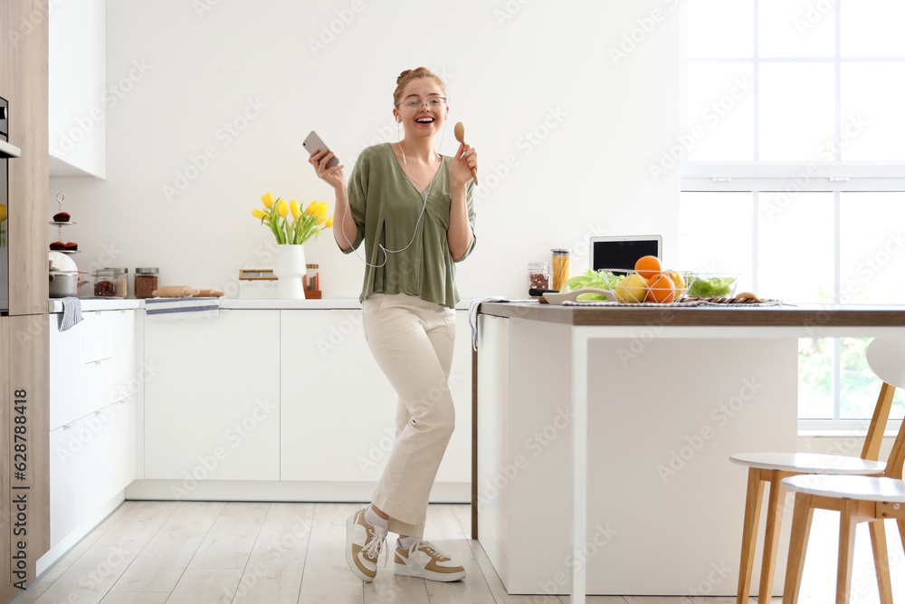 Pretty young woman listening to music, dancing and using wooden spoon as microphone in light kitchen