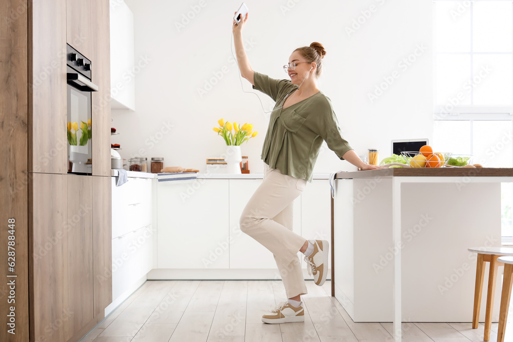 Happy young woman listening to music and dancing in light kitchen