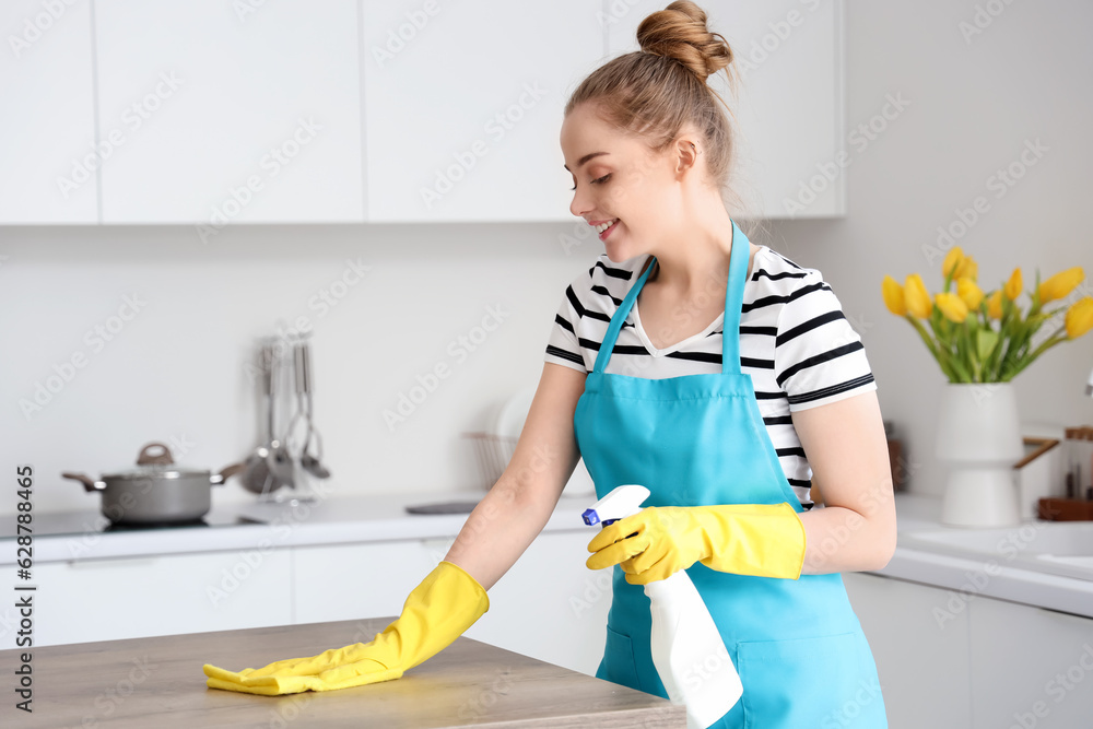 Happy young woman in yellow rubber gloves cleaning dining table with rag and detergent