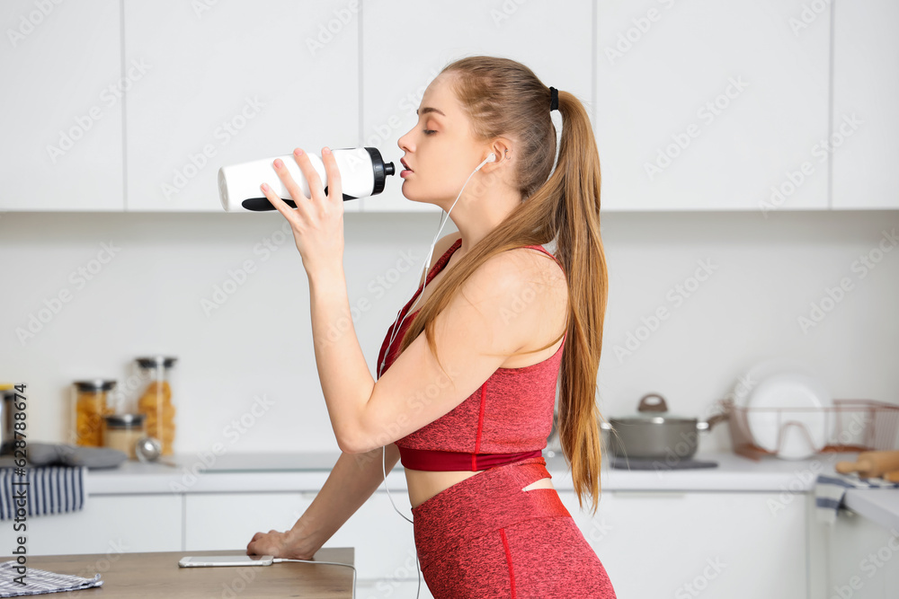 Sporty young woman drinking water from bottle in light kitchen