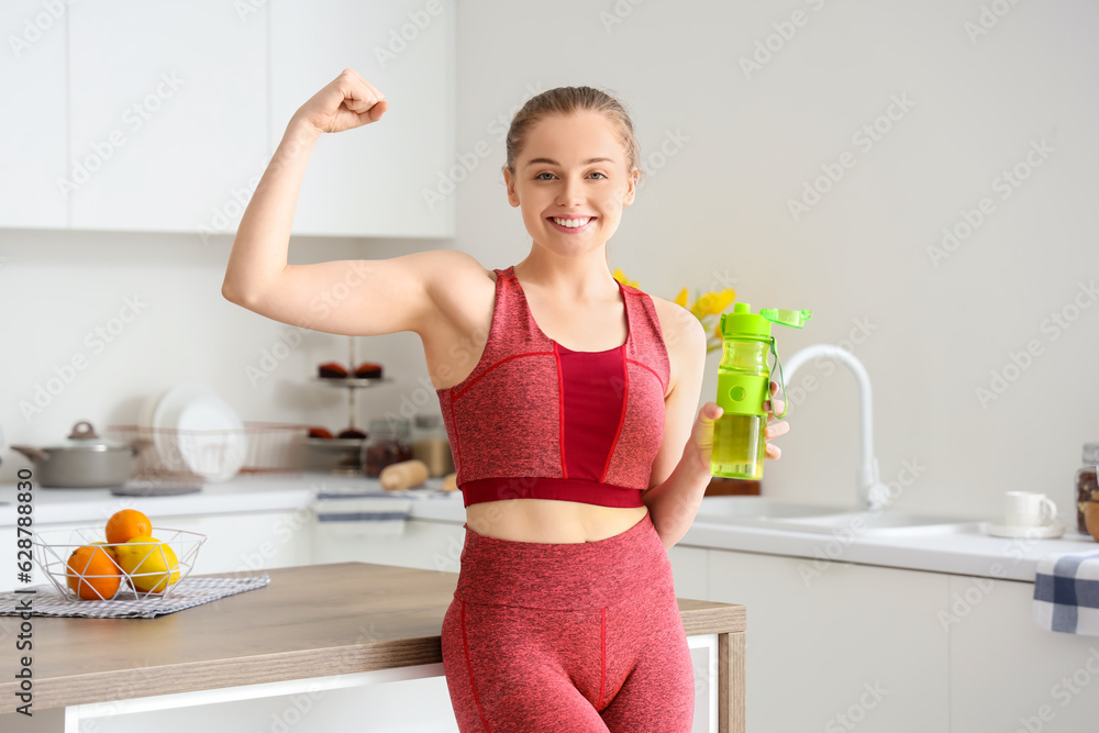 Sporty young woman with bottle of water flexing her arm muscles in light kitchen