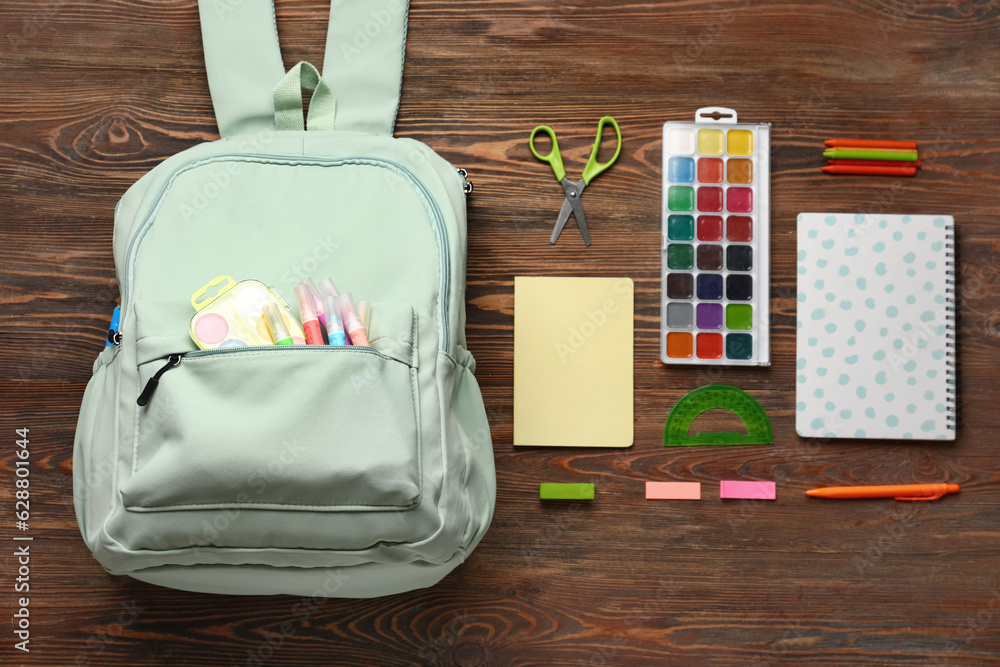 Green school backpack with watercolors, markers and notebooks on wooden background