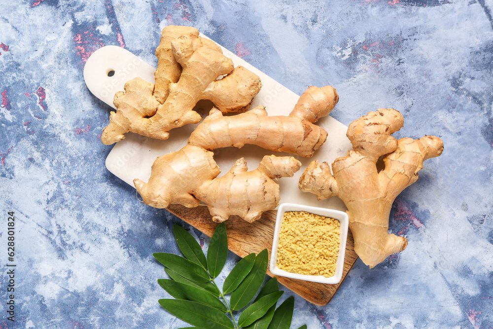 Board with fresh ginger roots, leaves and bowl of dried powder on blue background