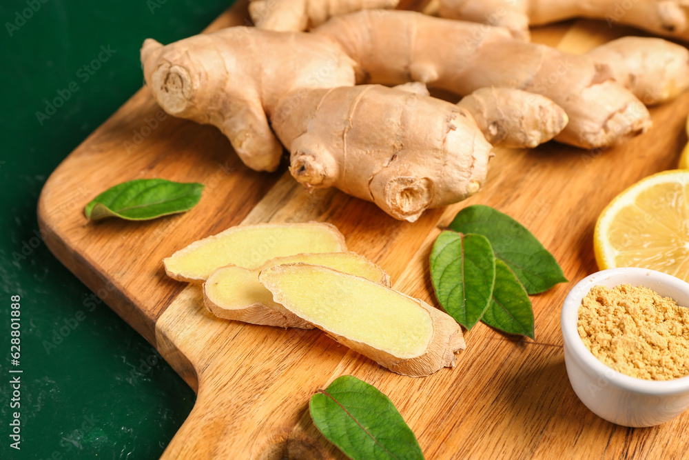 Wooden board with fresh ginger roots, sliced lemon and bowl of dried powder on green background