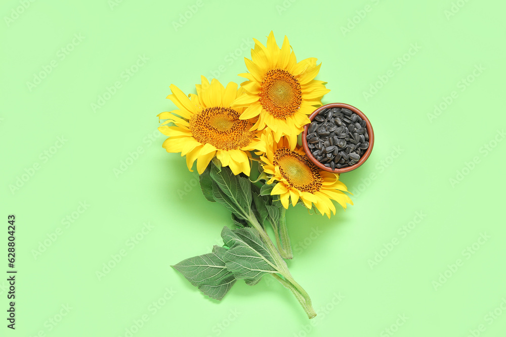 Beautiful sunflowers and bowl with seeds on green background