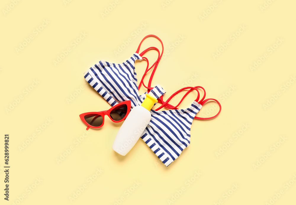 Sunglasses with bikini bra and bottle of sunscreen cream on pale yellow background