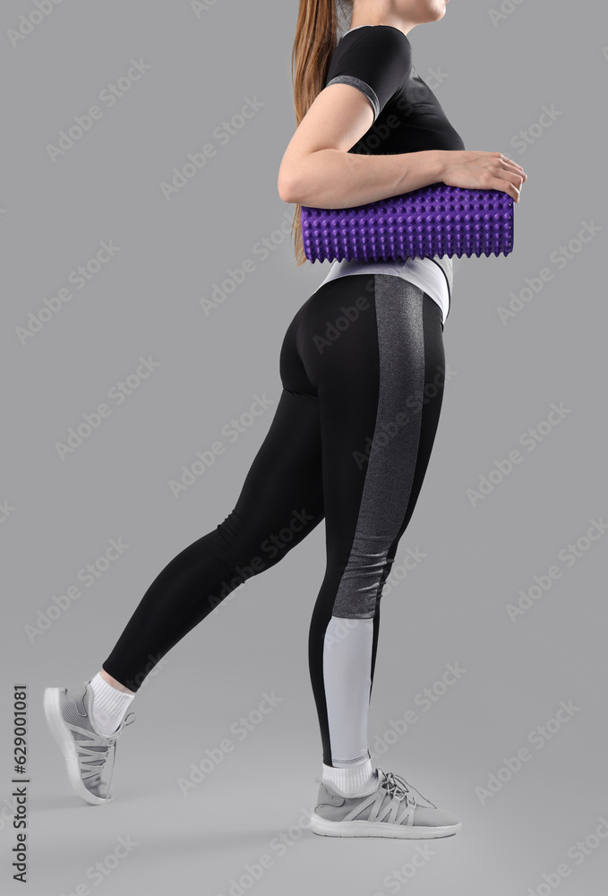 Young woman in sportswear and with foam roller on grey background