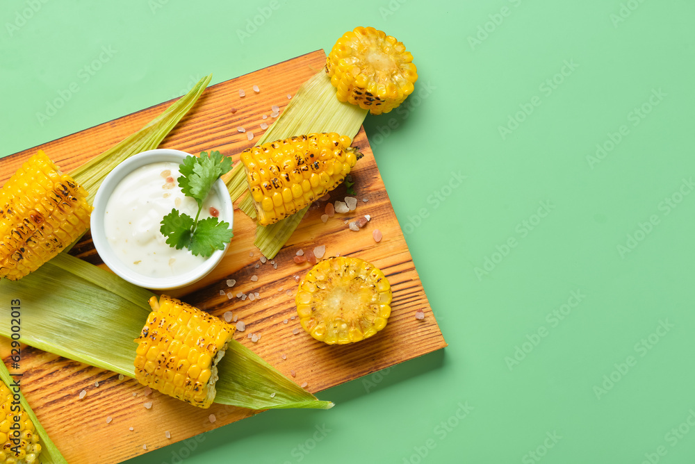 Wooden board with tasty grilled corn and sauce cobs on green background