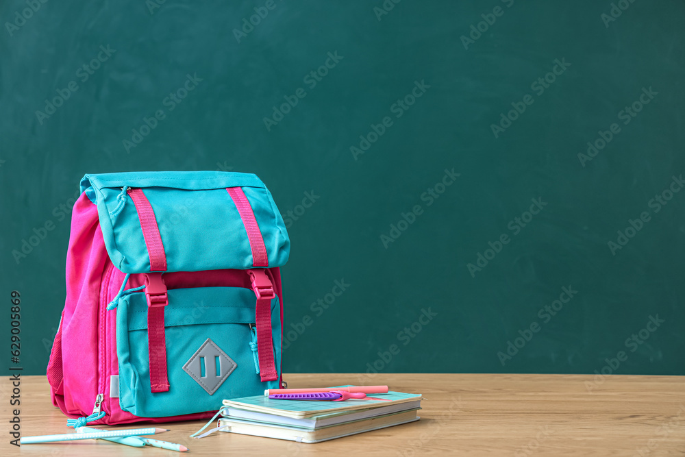 Colorful school backpack with stationery on brown wooden table near green chalkboard