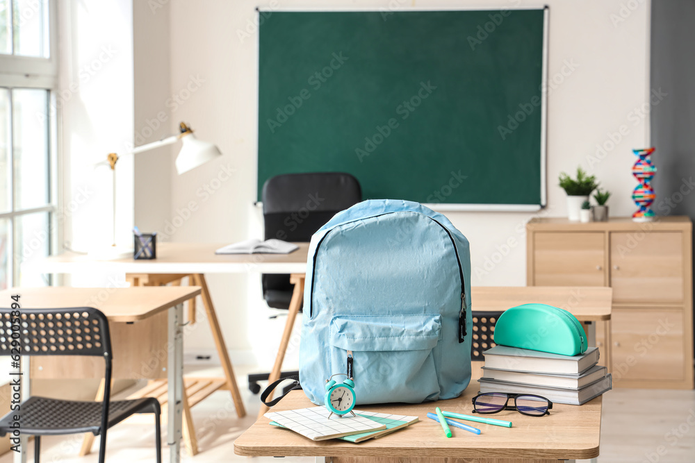 Blue school backpack with stationery, eyeglasses and alarm clock on desk in classroom