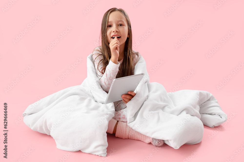 Little girl with blanket and tablet computer biting nails on pink background