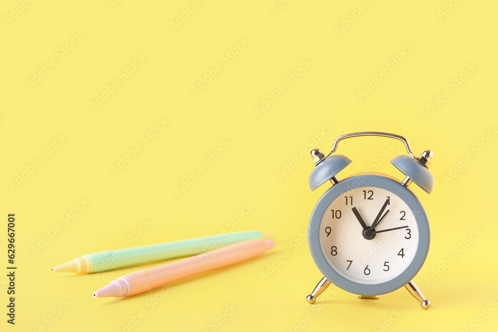 Alarm clock and pens on yellow background