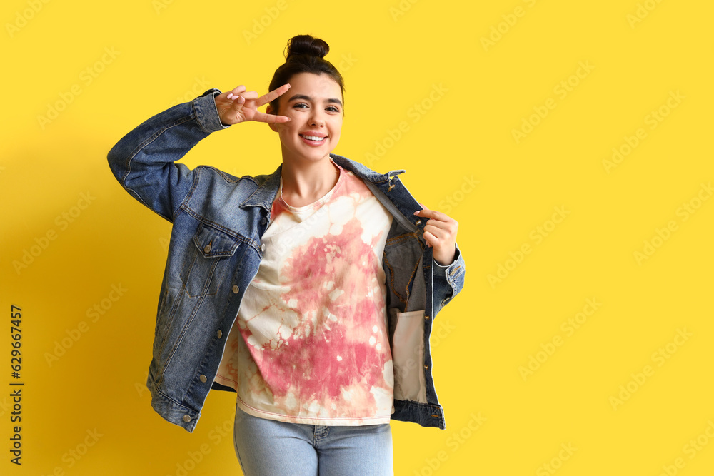 Young woman in tie-dye t-shirt showing victory gesture on yellow background