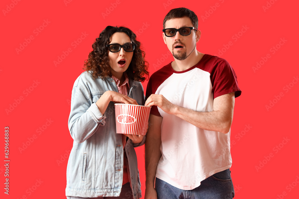 Shocked young couple in 3D glasses with popcorn on red background