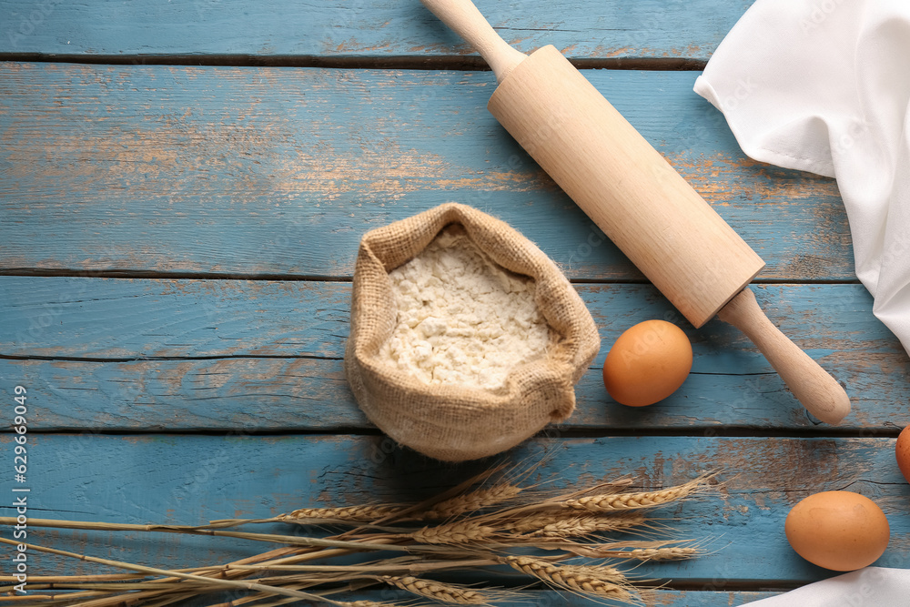 Bag of flour, eggs, wheat ears and rolling pin on blue wooden table