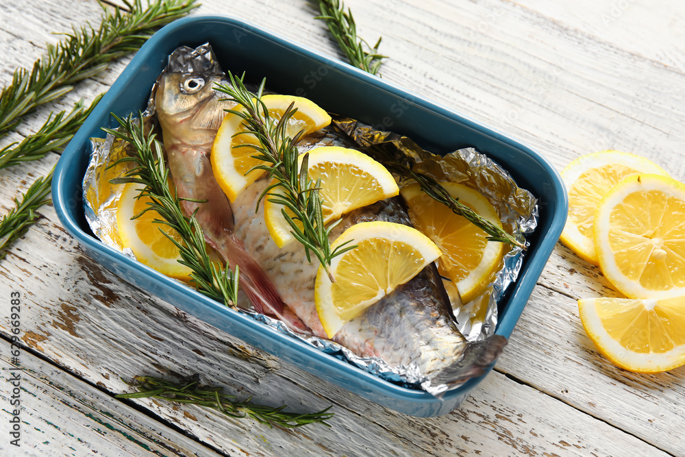 Baking dish with raw fish, lemon slices and rosemary on light wooden background