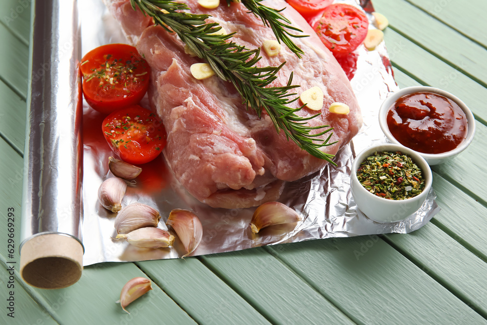 Aluminium foil roll with piece of raw meat, tomatoes, rosemary and spices on color wooden background