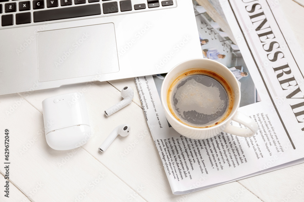 Cup of coffee with earphones, newspaper and laptop on white wooden background, closeup