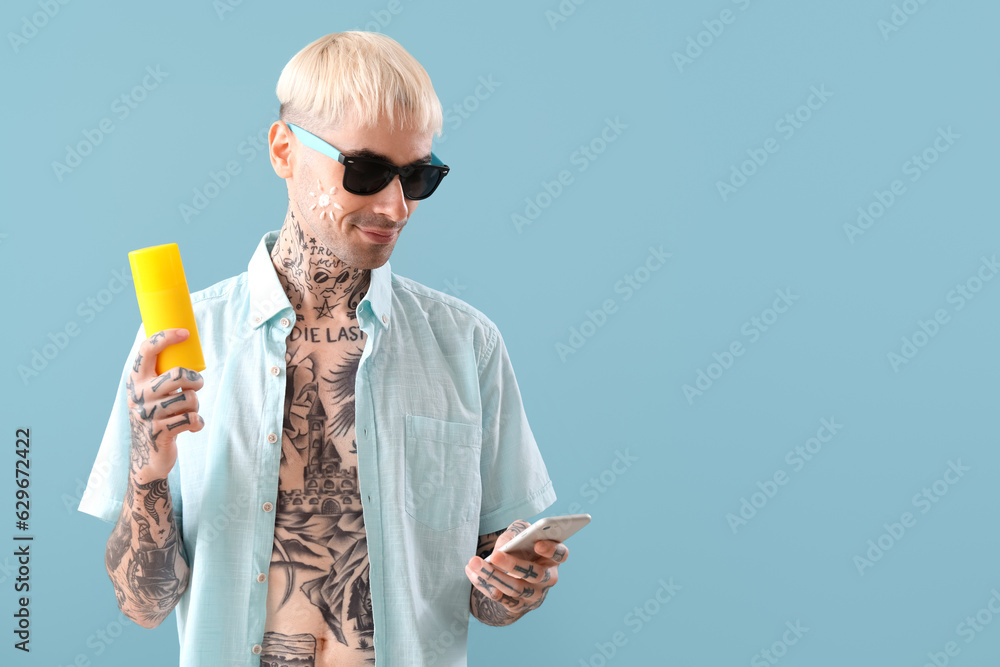 Tattooed man with phone and sunscreen cream on light blue background