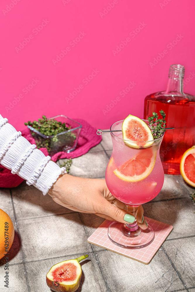 Woman holding glass of fresh summer cocktail with fig, grapefruit and thyme on tile table near pink 