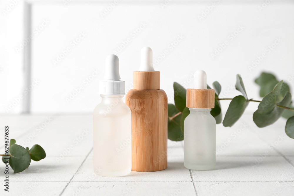 Bottles of cosmetic oil and eucalyptus branches on white tile