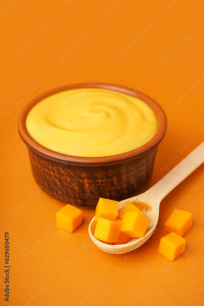 Bowl of tasty cheddar sauce and spoon with cheese cubes on orange background