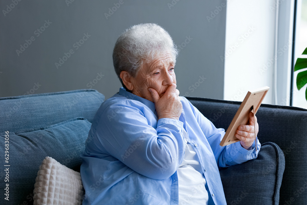 Sad senior woman with photo frame sitting on sofa at home