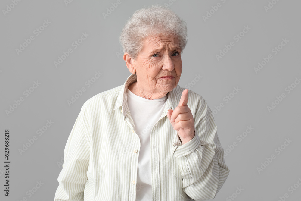 Upset senior woman with raised index finger on grey background