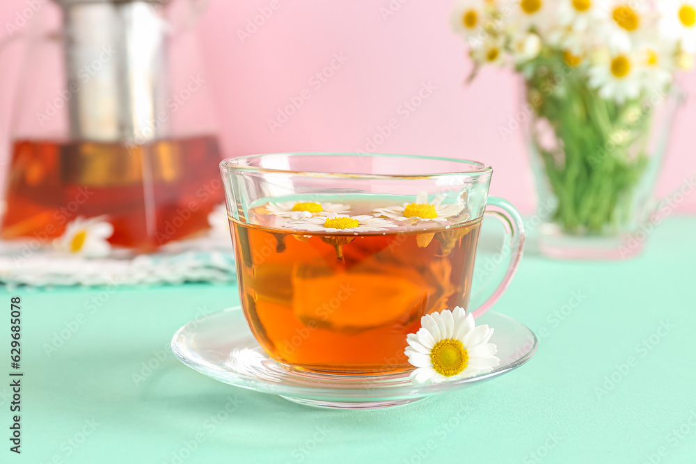 Teapot with cup of natural chamomile tea and flowers on turquoise table near pink wall