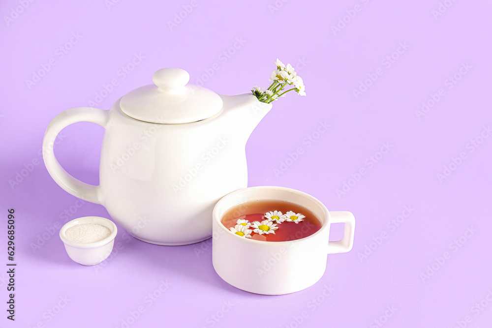 Teapot with cup of natural chamomile tea, sugar and flowers on lilac background