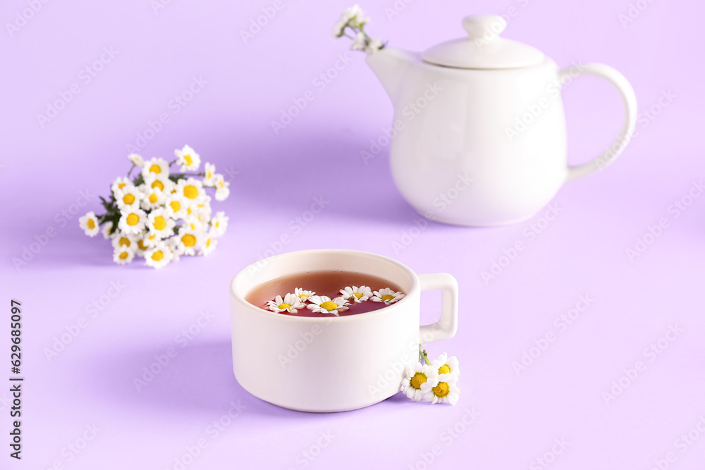 Teapot with cup of natural chamomile tea and flowers on lilac background