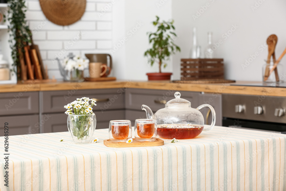 Wooden tray with teapot, cups of natural chamomile tea and flowers on table in kitchen